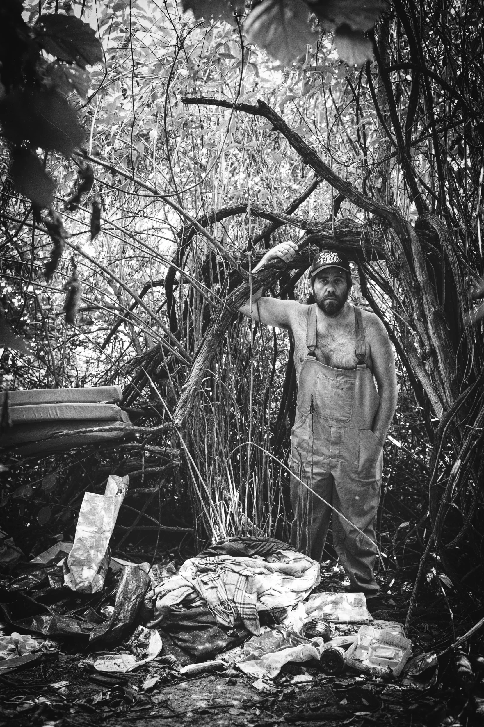 Full portrait of a man in overalls standing in a makeshift forest camp.